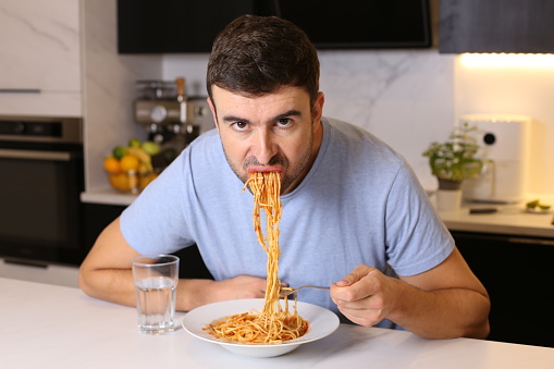 A man is trying to eat a classical spaghetti plate with tomato. He is hungry and is eating the pasta with bad manners .