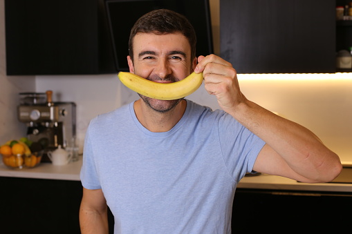 A cheerful man in his forties is covering his mouth with a delicious looking banana. He sits in his modern kitchen and has a playful mood.