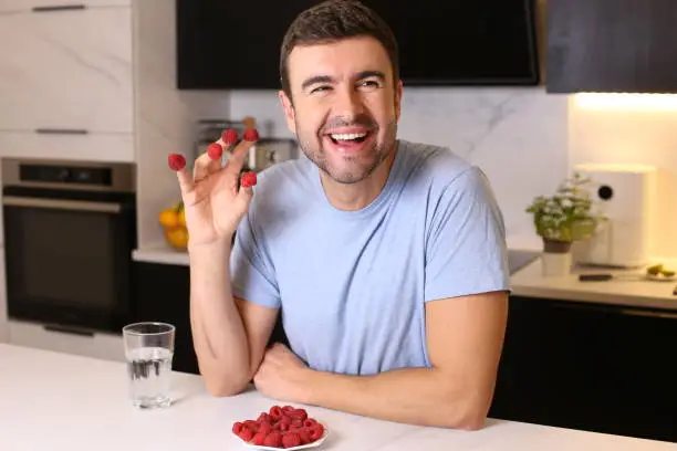 A playful man in his forties is enjoying a plate of fresh framboise. He sits in his modern looking kitchen and smiles.