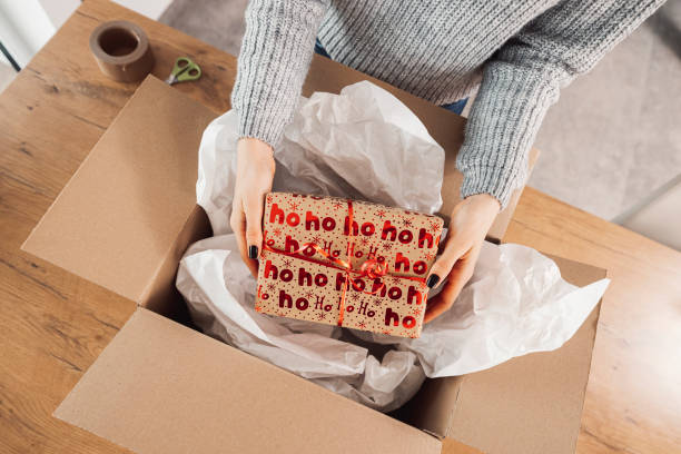 istock Top down view woman hands holding a Christmas present, putting it inside a cardboard box, ready to send in the mail 1449063568