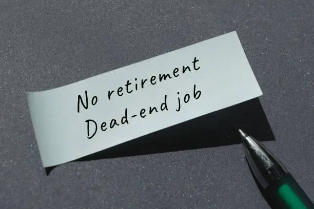 No retirement, dead-end job words written on paper note with pen. Handwritten note, caption, text. Top view, flat lay on dark grey paper background.