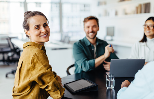 Female designer smiling at the camera while sitting at a table with her team. Business woman having a meeting with her colleagues in an office.