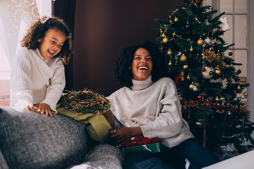 A cheerful Latin female and her cute little daughter laughing while using ornaments to prepare for Christmas And New Year celebration. They are sitting on the sofa.