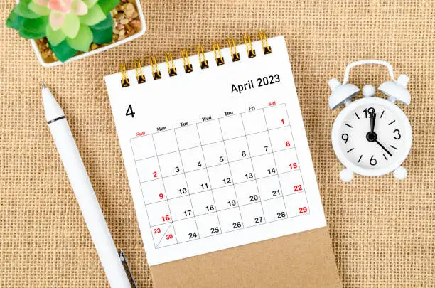 Photo of The April 2023 Monthly desk calendar for the organizer to plan 2023 year with alarm clock and pen on sack background.