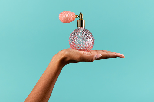 Crop unrecognizable African American female demonstrating pink crystal vintage bottle with spray of perfume on turquoise background