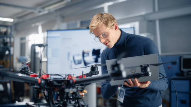 istock Male Developer Works on Engineering Project, Codes on Laptop and Looks at Drone in Modern Office. Empowered Person Creating Futuristic Unmanned Aerial Vehicles for Delivery Concept. 1449083889