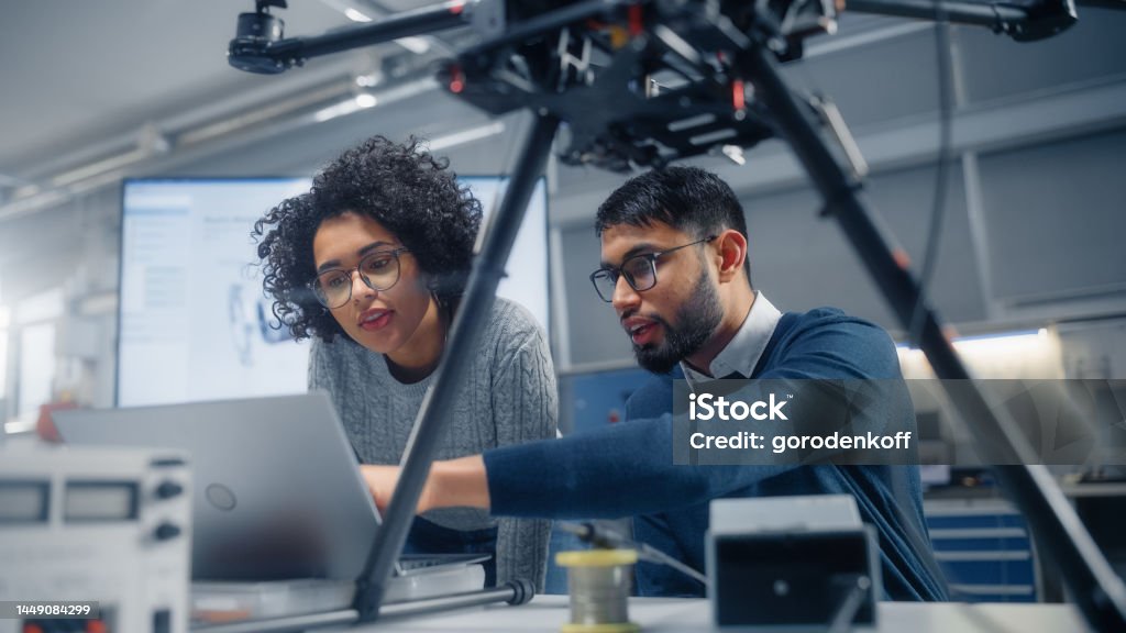 Black Male Engineer Points at Laptop Screen and Talks. Brainstorming New Drone Design With his Female Colleague. Innovative Technologies in Unmanned Aerial Vehicle Design Concept. Engineer Stock Photo