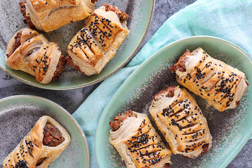 Stock photo showing close-up, elevated view of freshly baked, homemade sausage rolls topped with black sesame seeds, on green plates besides turquoise muslin against a mottled, grey background.