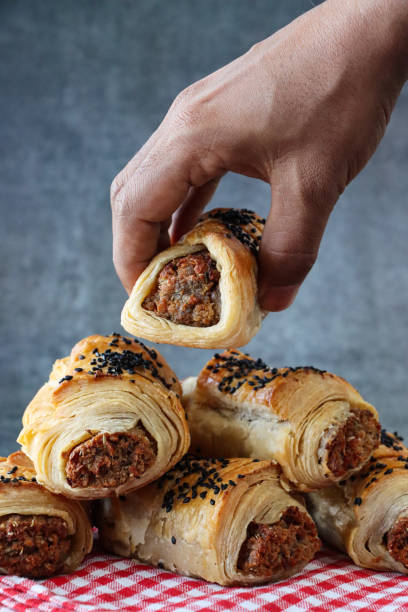 Close-up image of unrecognisable person selecting sausage roll, stacked heap of baked, homemade sausage rolls topped with black sesame seeds, on red and white gingham checked tea towel, mottled, grey background, focus on foreground stock photo