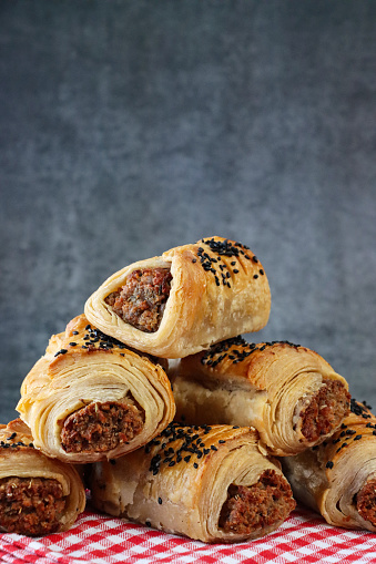 Stock photo showing close-up view of a heap of freshly baked, homemade sausage rolls topped with black sesame seeds, on a red and white gingham checked tea towel against a mottled, grey background.