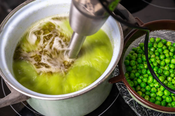 Stick blender blends pea soup in pot on hob, raw pea. stock photo