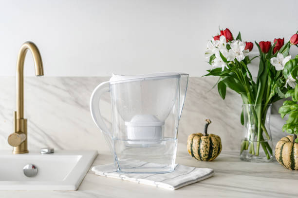water filter jug with napkin on kitchen table water purification in pitcher with filter, blossom flowers in glass vase and pumpkin on countertop, sink and golden faucet at home kitchen Water Filter stock pictures, royalty-free photos & images