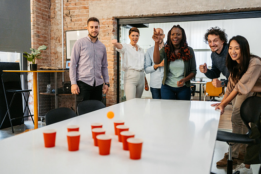 Colleagues taking a break in the office, playing beer pong with empty cups on the office table.