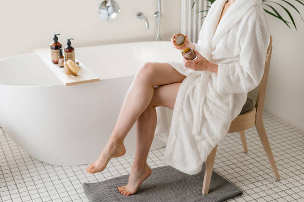 Woman in terry robe holding glass jar with face cream Woman in terry robe holding glass jar with face cream. Lady sitting on chair close to tub with cosmetics on shelf. Female spend morning in bathroom, making pampering and moisturizing procedure at home woman legs open stock pictures, royalty-free photos & images