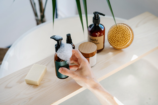 High angle view of woman hand choose cosmetics product for everyday skincare routine. Female hold glass bottle with dispenser over wooden shelf in bathroom. Self care, moisturizing, pampering concept