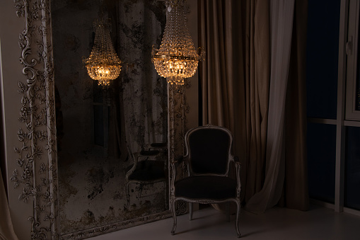 A crystal old chandelier hangs near the mirror and there is a chair next to the house in the evening near the window, apartment interior