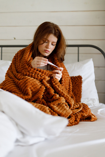 Woman at home measures high temperature, wrapped in a warm blanket on the bed, fever. Disease.