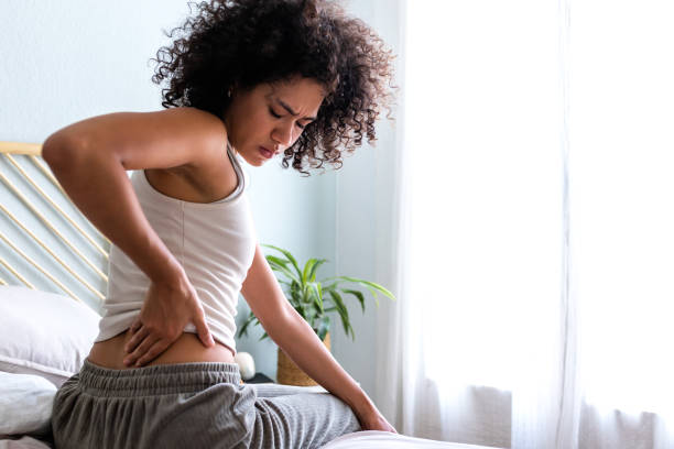 rear view of young woman touching lumbar feeling back pain. female sitting on bed with back ache. copy space. - dor de costas imagens e fotografias de stock