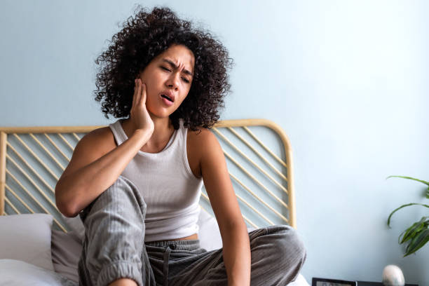 Young latina female with curly hair touching cheek in pain having toothache sitting on bed in cozy bedroom at home. Young latina female with curly hair touching cheek in pain having toothache sitting on bed in cozy bedroom at home. Body concept. jaw pain stock pictures, royalty-free photos & images