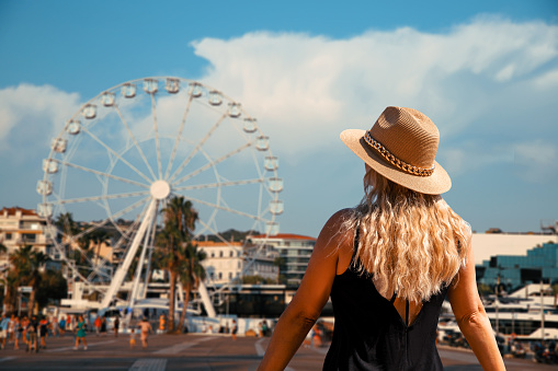 Rear view of Blonde Woman with sun hat Discover the city of Cannes, France,  looking at city Ferris Wheel
