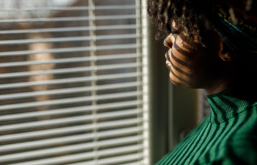 Portrait of serious young Black woman standing by the window, looking outside, as if she is waiting for someone. Window blinds throwing interesting shade over her face and chest.