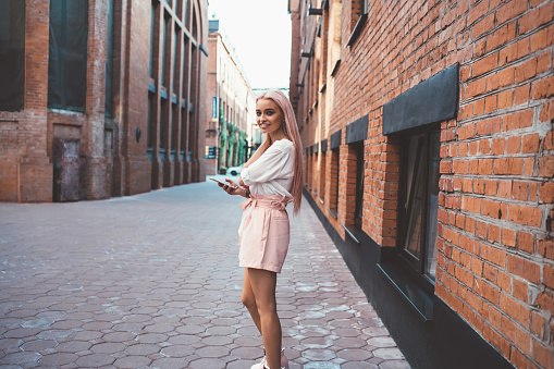 Carefree female youngster smiling at urbanity street enjoying leisure time for web blogging via cellular device, happy adolescent woman with pink hair using smartphone while walking in city