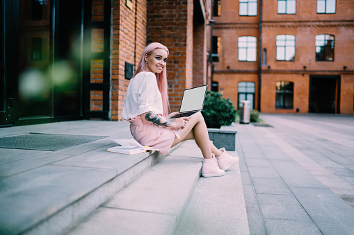 Cheerful female student sitting at city urbanity with mockup laptop computer and education textbook for doing distance e learning, happy Caucasian freelance journalist with blank netbook smiling