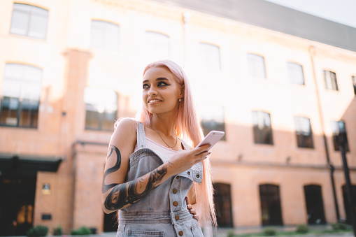 Cheerful female blogger with tattooed hand looking away and smiling during leisure time in city, pretty Caucasian hipster girl with pink hair holding modern smartphone connected to 4g wireless