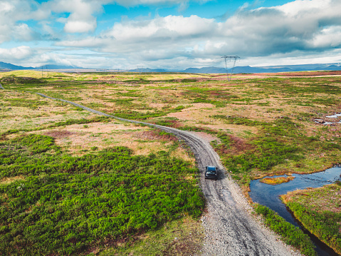 Remote dirt road somewhere in Iceland mainland, surrounded by vibrant green bushes and volcanic lands. A car driving alone on the gravel road.