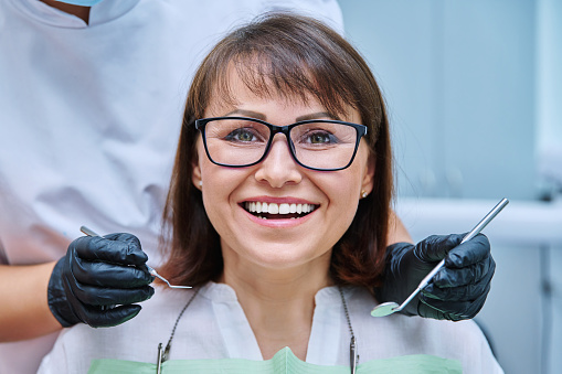 Close-up face of smiling mature woman with teeth in dentistry with hands with dentist examination tools. Treatment, prosthetics, implantation, whitening, hygiene, dental health care concept