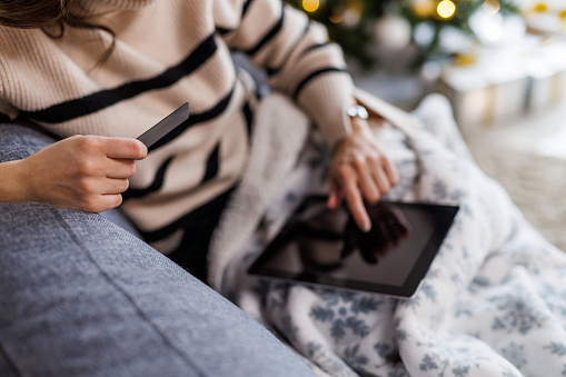 Cut out of contented young woman lounging on the sofa in her living room, holding her credit card and doing online Christmas shopping via digital tablet.