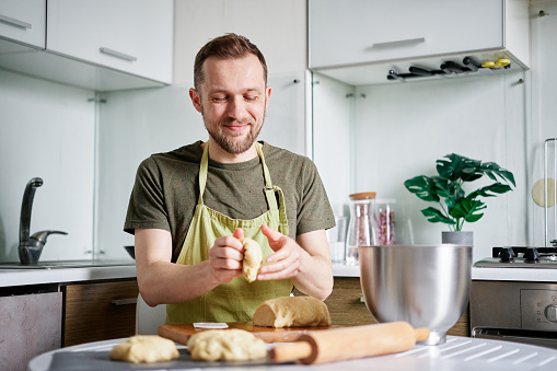 Attractive caucasian bearded chef baker in green apron working with dough on wooden cutting board with rolling pin on foreground. Working at home kitchen concept, homemade baking. High quality photo