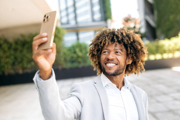 istock Selfie photo concept. Positive handsome curly haired hispanic man standing outdoors, taking a selfie on his smartphone, mixed race male blogger having live streaming on social network, smiling 1449140610