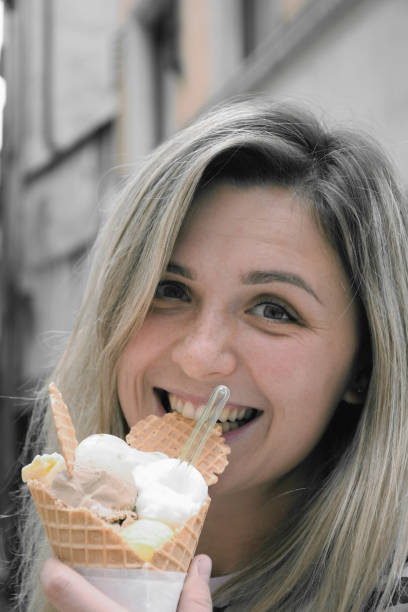 A young woman is eating an ice cream cone and smiling. The background is blurred