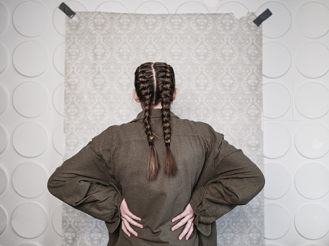 Studio Portrait of a girl actor in costume from the back. She is dressed in raggedy clothes of a poor person and has braided hair. Close up image in front of the damask pattern backdrop.