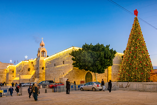 People walk on Manger Square in downtown Bethlehem, Palestine, Israel in the evening, during the Christmas Holidays, with the Church of the Nativity in the background.
