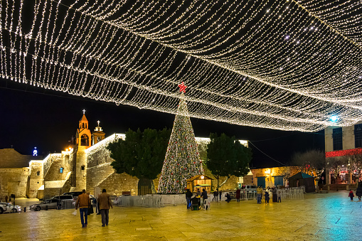 People walk on Manger Square in downtown Bethlehem, Palestine, Israel in the evening, during the Christmas Holidays, with the Church of the Nativity in the background.