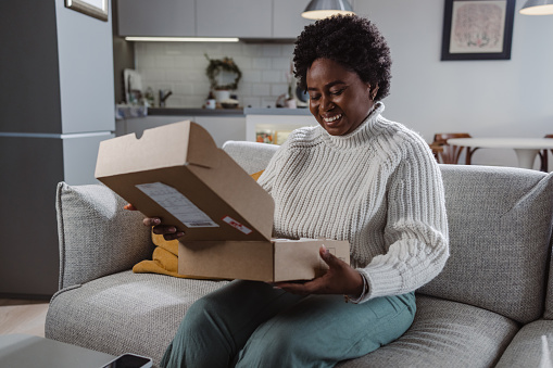 African-American woman sitting on the sofa in the living room and unpacking a cardboard box