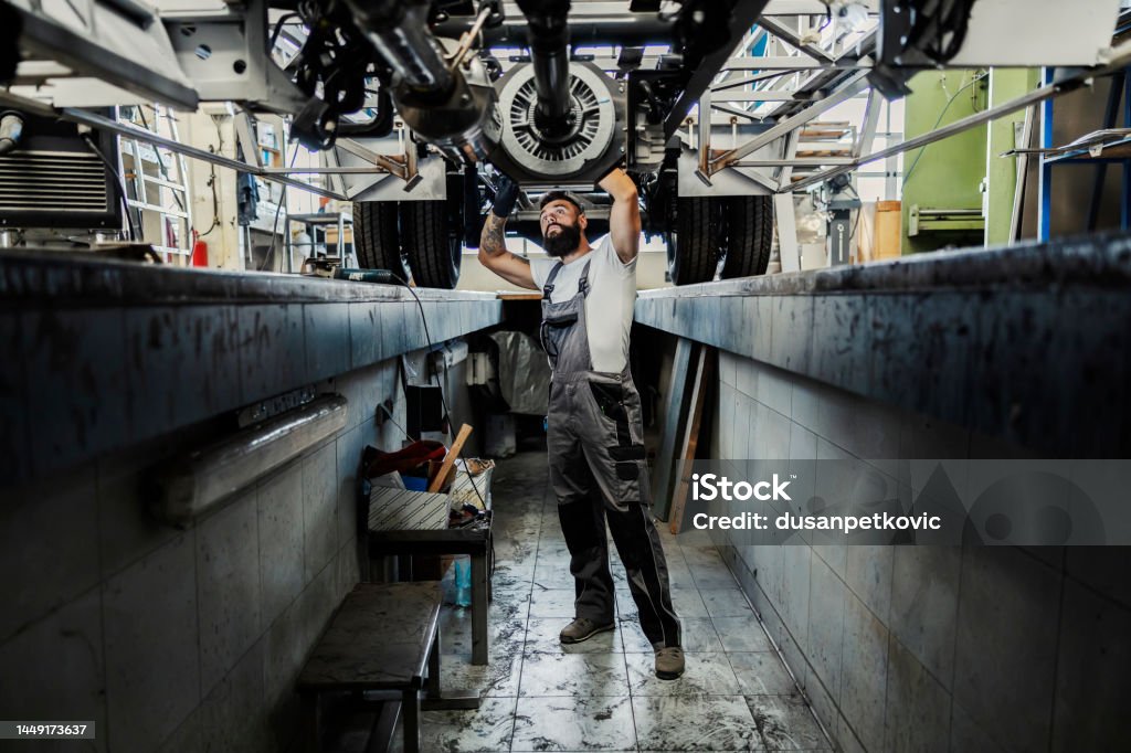 An auto-mechanic in a pit looking at broken vehicle and trying to fix it. A mechanic in a workshop fixing vehicle. Automobile Industry Stock Photo