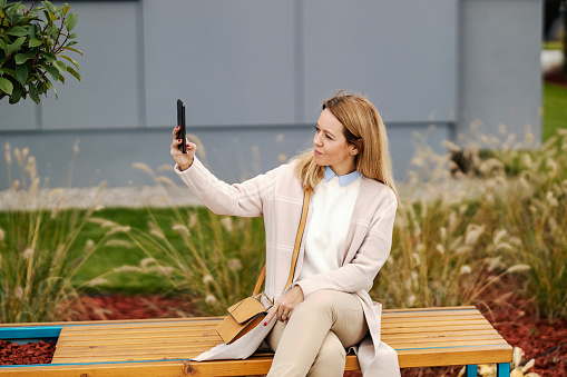 A trendy middle-aged woman is sitting on a bench in city park and taking self portraits for online photo messaging.