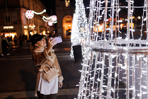 One smiling young woman taking selfie on mobile phone in Christmas decorated city at night