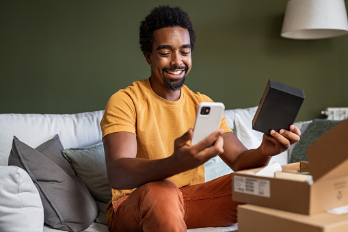 Young man sitting at the table surrounded by cardboard boxes. African-American man is unboxing delivery package in the living room. His new mobile phone has arrived.