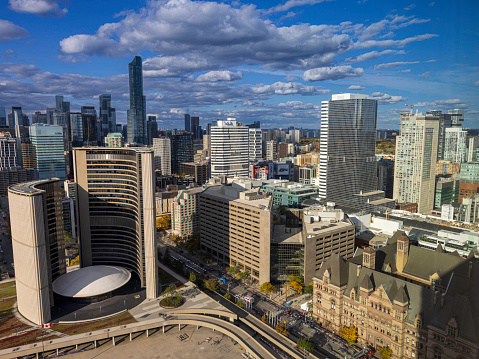 view to the Old City Hall of Toronto, modern buildings behind, with cloudy sky. the Arch and Fountain at Nathan Phillips Square in front.