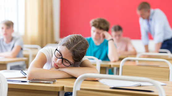 Exhausted teen girl sleeping at desk in classroom during lesson with blurred classmates in background