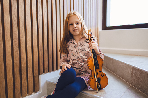 Young girl holding a violin in a portrait photo while sitting on the stairs, at home.