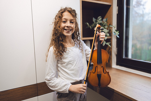 Young girl smiling while standing and holding a violin in her room, at home.