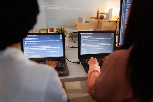 Rear view of two female programmers sitting at the table and typing on their laptops while developing new software