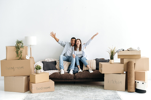 Excited happy multiracial young couple in love, sitting on sofa in living room of their new home between cardboard boxes, they invested in real estate, happy with purchase, looking at camera, smiling