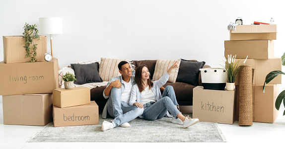 Excited joyful mixed race couple in love, sitting on the floor in the living room in their new home, planning interior design of their new apartment, cardboard boxes with things are standing nearby