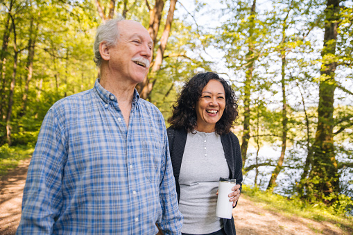 Grey-haired man with a moustache and a Latina woman slowly strolling by a pond in a city park. The lady holding a flask and laughing heartily. Waist up image, looking away.